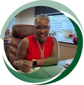 A smiling woman with short gray hair and red glasses sits at a desk in an office, writing in a notebook with a green pen. She’s wearing a sleeveless red dress, a beaded necklace, and an Apple Watch, with a window view of buildings and greenery in the background.